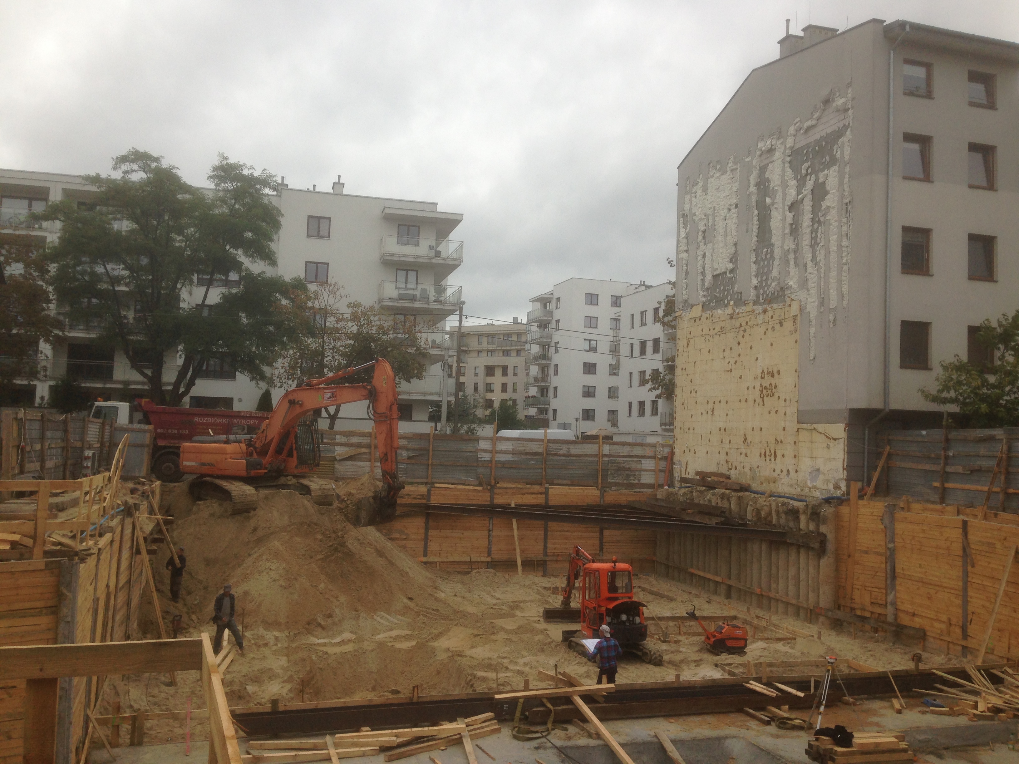 Pomarańczowa koparka pracuje przy wykopie zabezpieczonym drewnianą konstrukcją, w tle budynki mieszkalne w pochmurny dzień.