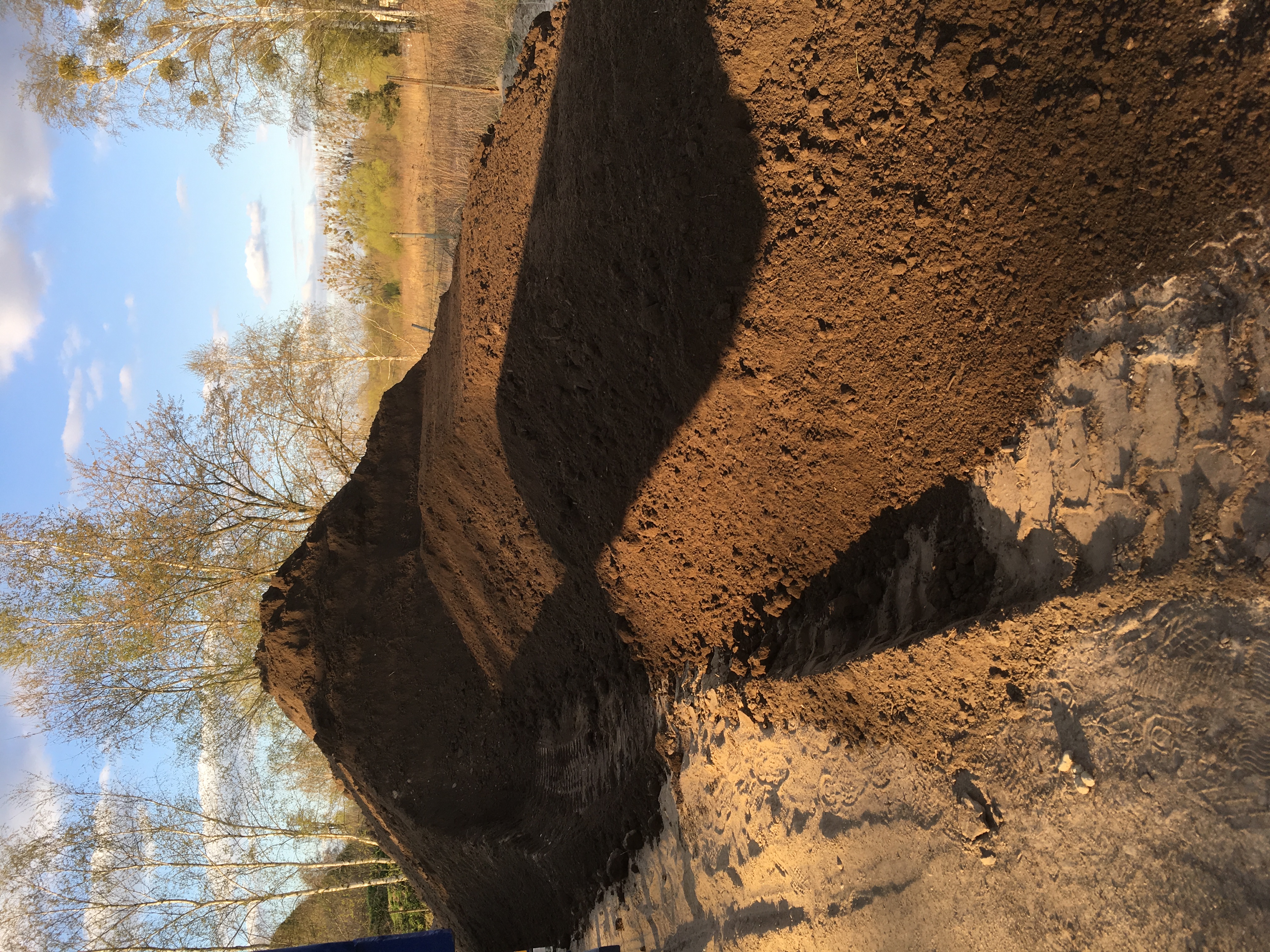 Wysoka pryzma brązowej ziemi ogrodowej, częściowo zacieniona, z widocznymi śladami opon i drzewami w tle pod błękitnym niebem.