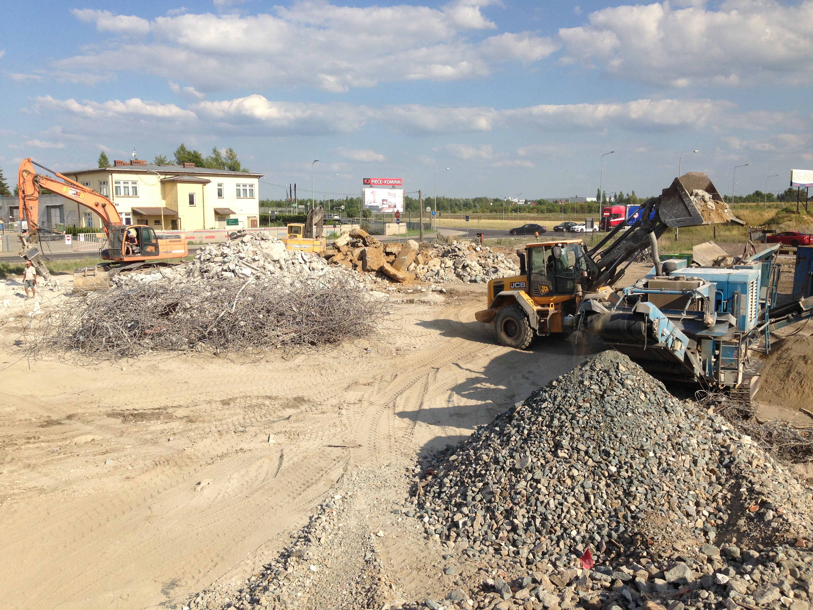 Prace rozbiórkowe z użyciem koparki i przesiewacza gruzu na placu budowy, widoczne zbrojenia i stosy gruzu.
