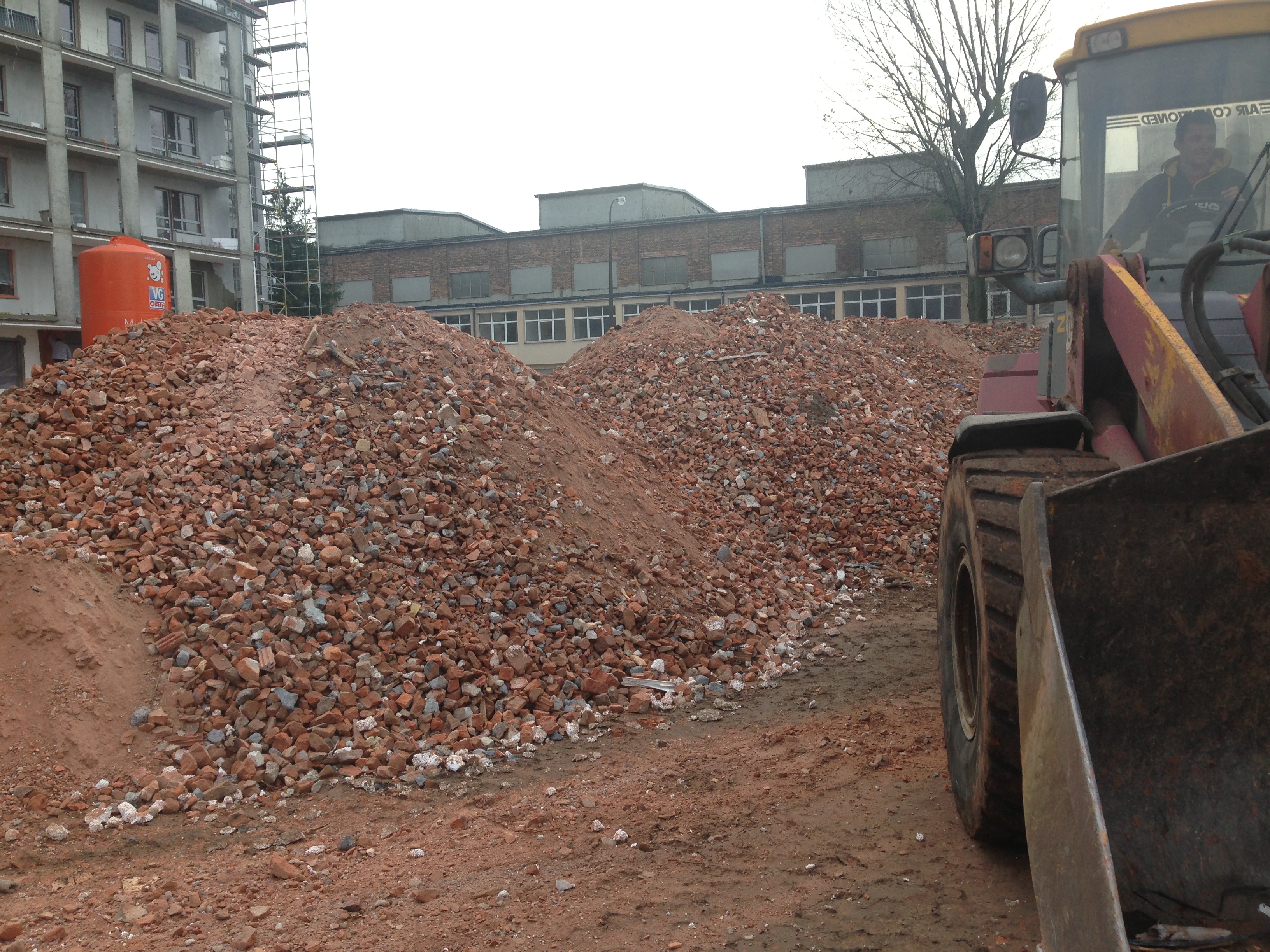 Stos gruzu ceglanego i ziemi z fragmentem żółto-czerwonej ładowarki na pierwszym planie, w tle budynek mieszkalny w trakcie budowy i budynek przemysłowy.