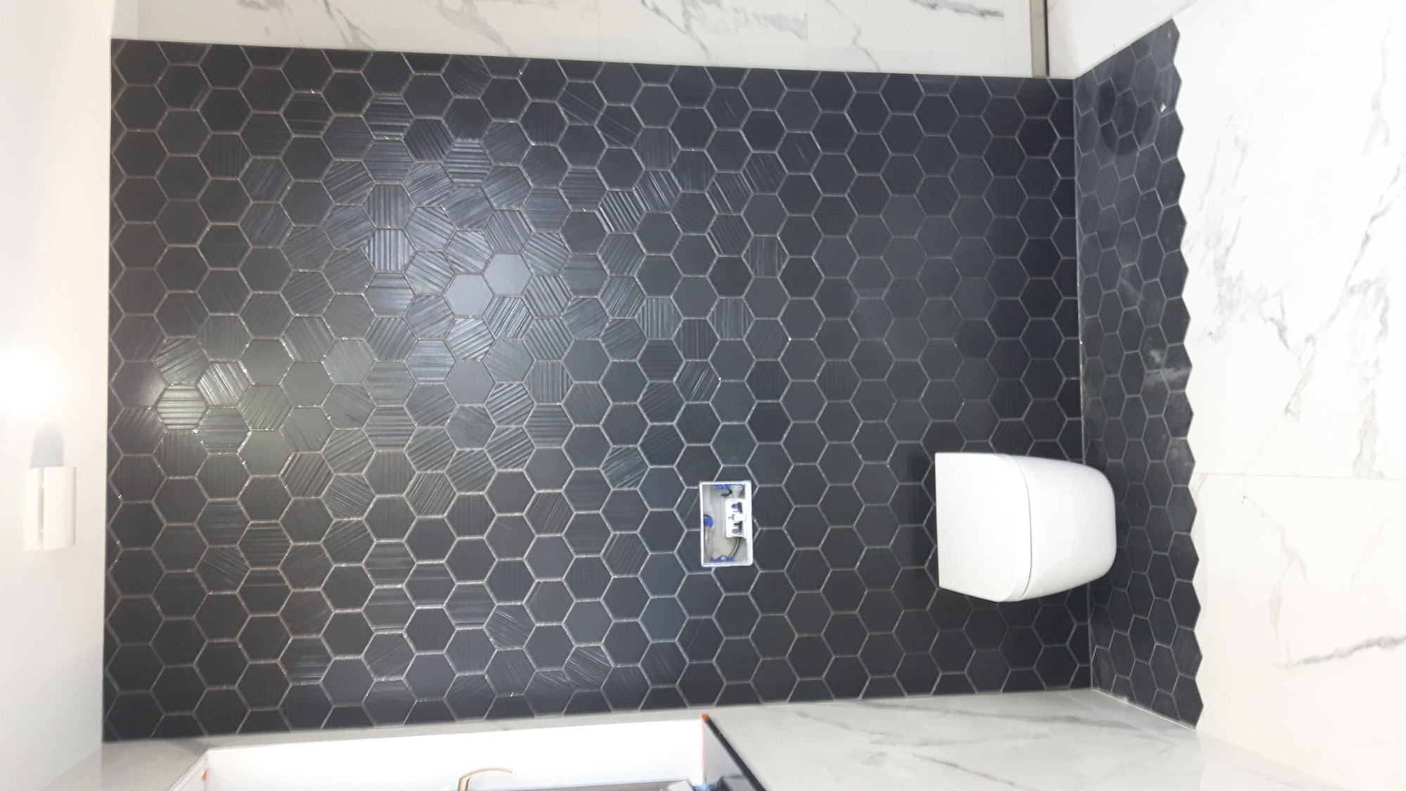 Ściana w łazience wyłożona czarnymi, heksagonalnymi płytkami z subtelnym wzorem, kontrastująca z białymi płytkami imitującymi marmur na sąsiedniej ścianie; widoczna biała toaleta podwieszana...