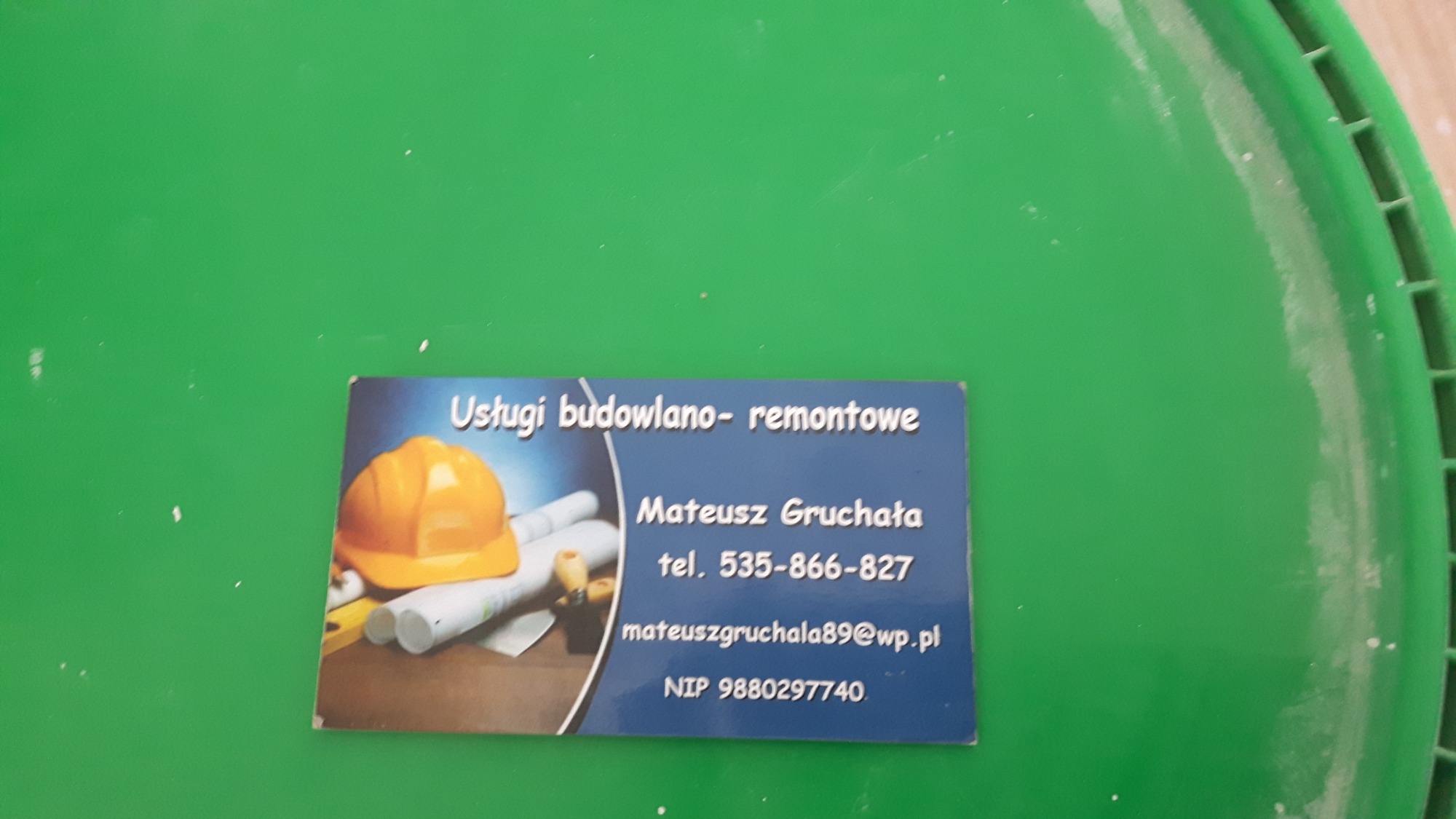 Wizytówka firmy budowlano-remontowej Mateusz Gruchała, leżąca na pokrywie wiadra z farbą. Na wizytówce grafika z kaskiem budowlanym i planami.