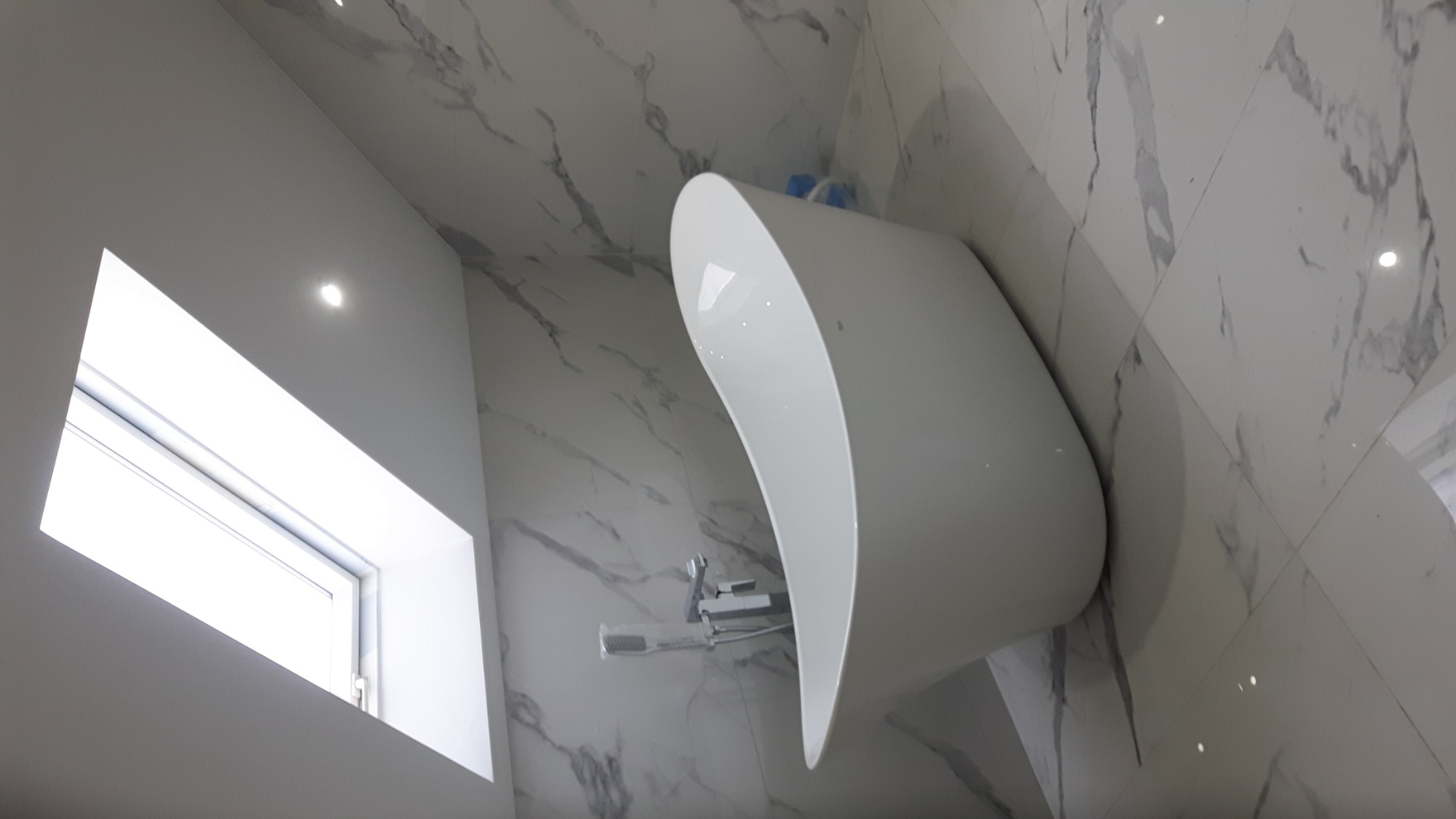 Nowoczesna, wolnostojąca wanna o organicznym kształcie, zamontowana bateria ścienna, marmurowe płytki na ścianach, jasne, naturalne światło wpadające przez okno.