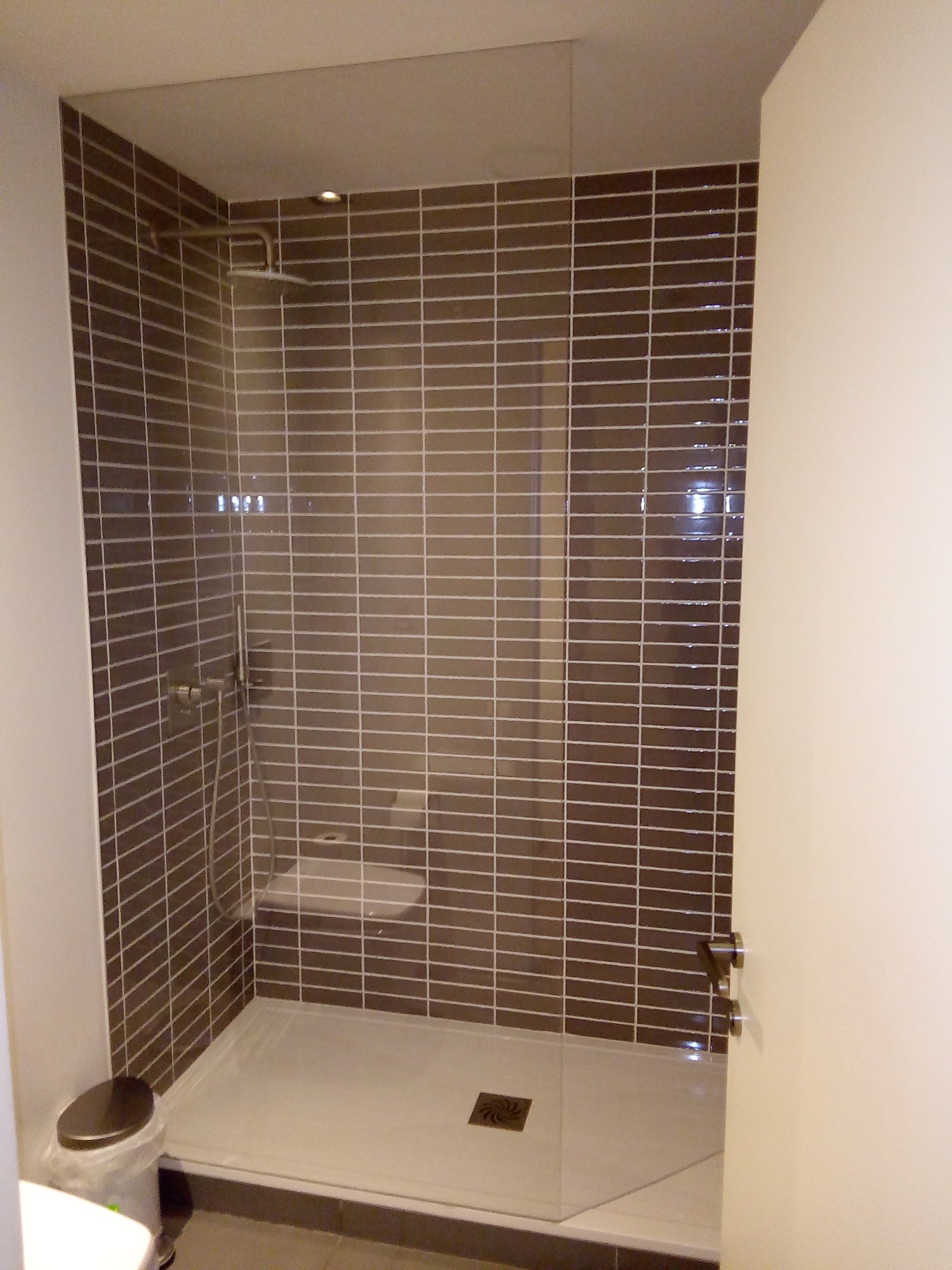 Nowoczesna kabina prysznicowa z ciemnobrązowymi, prostokątnymi płytkami ułożonymi poziomo, biały brodzik z odpływem w rogu, szklana ściana prysznicowa z chromowanym uchwytem, widoczny kosz na śmieci.