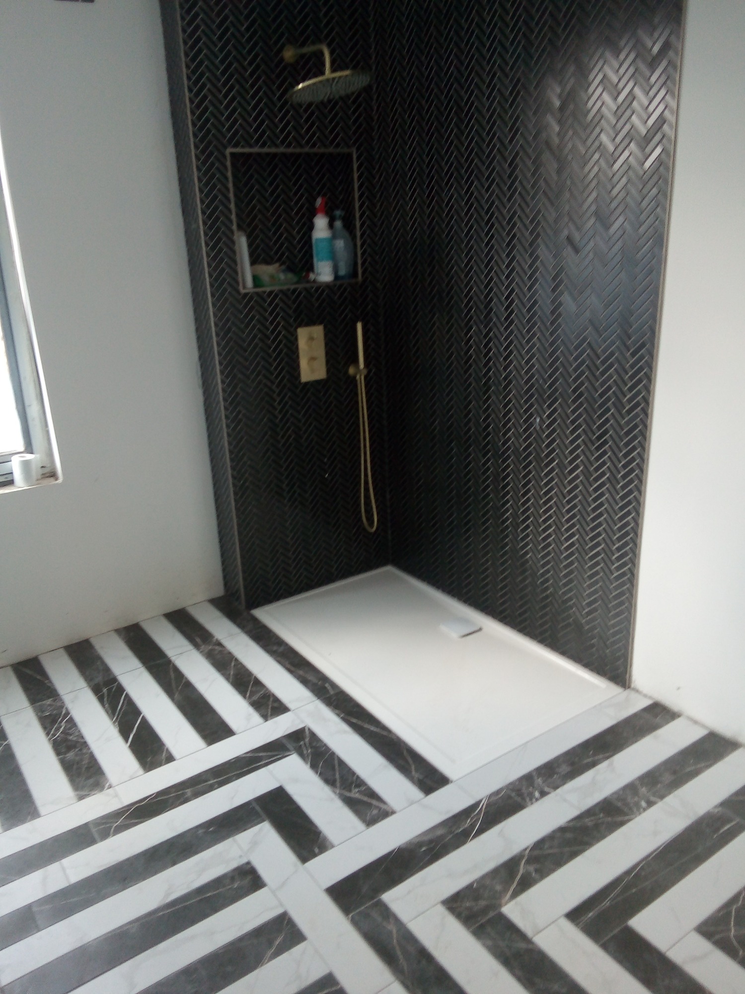 Wykończona łazienka z kabiną prysznicową wyłożoną czarną, wąską mozaiką w jodełkę, biały brodzik, złota armatura, półka z kosmetykami i podłoga w biało-czarne pasy.