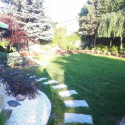 Zadbany ogród z równo przystrzyżonym trawnikiem, ścieżką z płyt kamiennych, rabatami z białym żwirem i różnorodną roślinnością, w tym drzewami iglastymi i liściastymi.