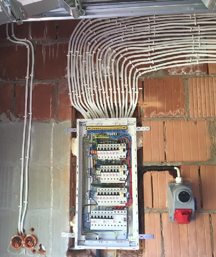 Rozdzielnica elektryczna z licznymi przewodami w białych peszlach, zamontowana na surowej ścianie z cegły w budynku w Koninie. Widoczny również gniazdo siłowe.