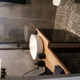 Nowoczesna łazienka z okrągłą umywalką nablatową, drewnianym blatem i czarnymi akcesoriami na tle wzorzystych płytek.