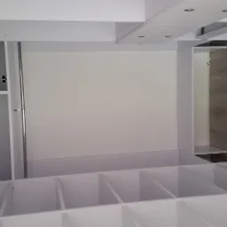 Wnętrze szafy na wymiar w minimalistycznym stylu, z białymi półkami i drążkiem na ubrania, widoczne oświetlenie sufitowe punktowe, jasne panele podłogowe.