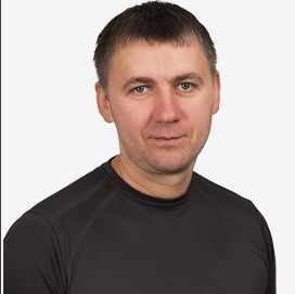 Portret mężczyzny w czarnej koszulce z krótkim rękawem, z krótkimi ciemnymi włosami i niebieskimi oczami, na jasnym tle.