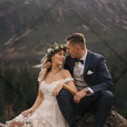 Para młoda w strojach ślubnych siedzi na skale z widokiem na góry; panna młoda ma wianek na głowie, a pan młody ma na sobie granatowy garnitur w kratę.