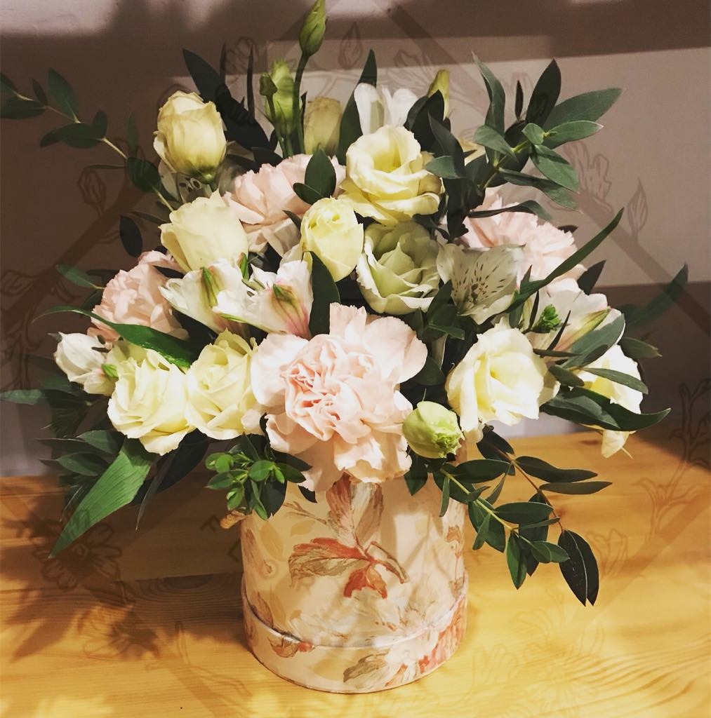 Kompozycja kwiatowa w okrągłym pudełku z motywem liści, zawierająca goździki, eustomy i alstromerie w odcieniach bieli, różu i zieleni, z dodatkiem zielonych gałązek eukaliptusa, ustawiona...