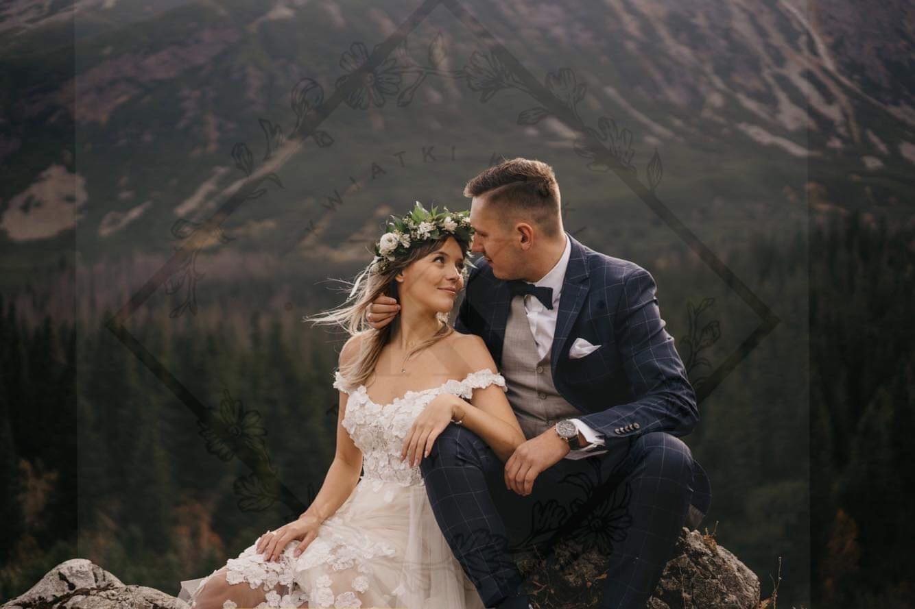 Para młoda w strojach ślubnych siedzi na skale z widokiem na góry; panna młoda ma wianek na głowie, a pan młody ma na sobie granatowy garnitur w kratę.