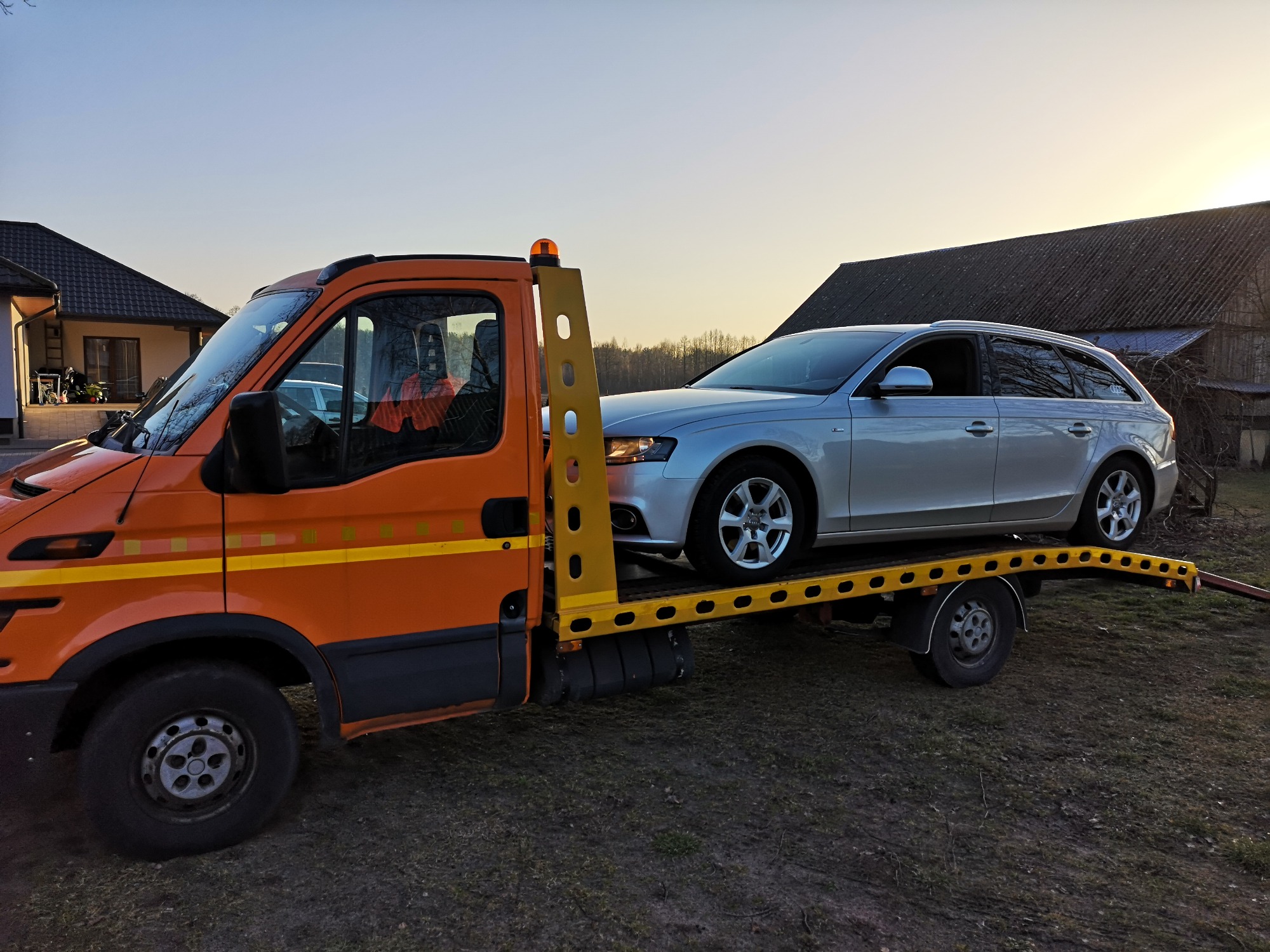 Pomarańczowa laweta z srebrnym kombi Audi na platformie, zaparkowana na trawiastym terenie przed wiejskim domem i stodołą o zmierzchu.