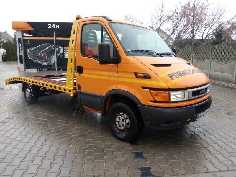 Pomarańczowa laweta Iveco z napisem 'POMOC DROGOWA' na masce, zaparkowana na mokrym placu przed myjnią samochodową.