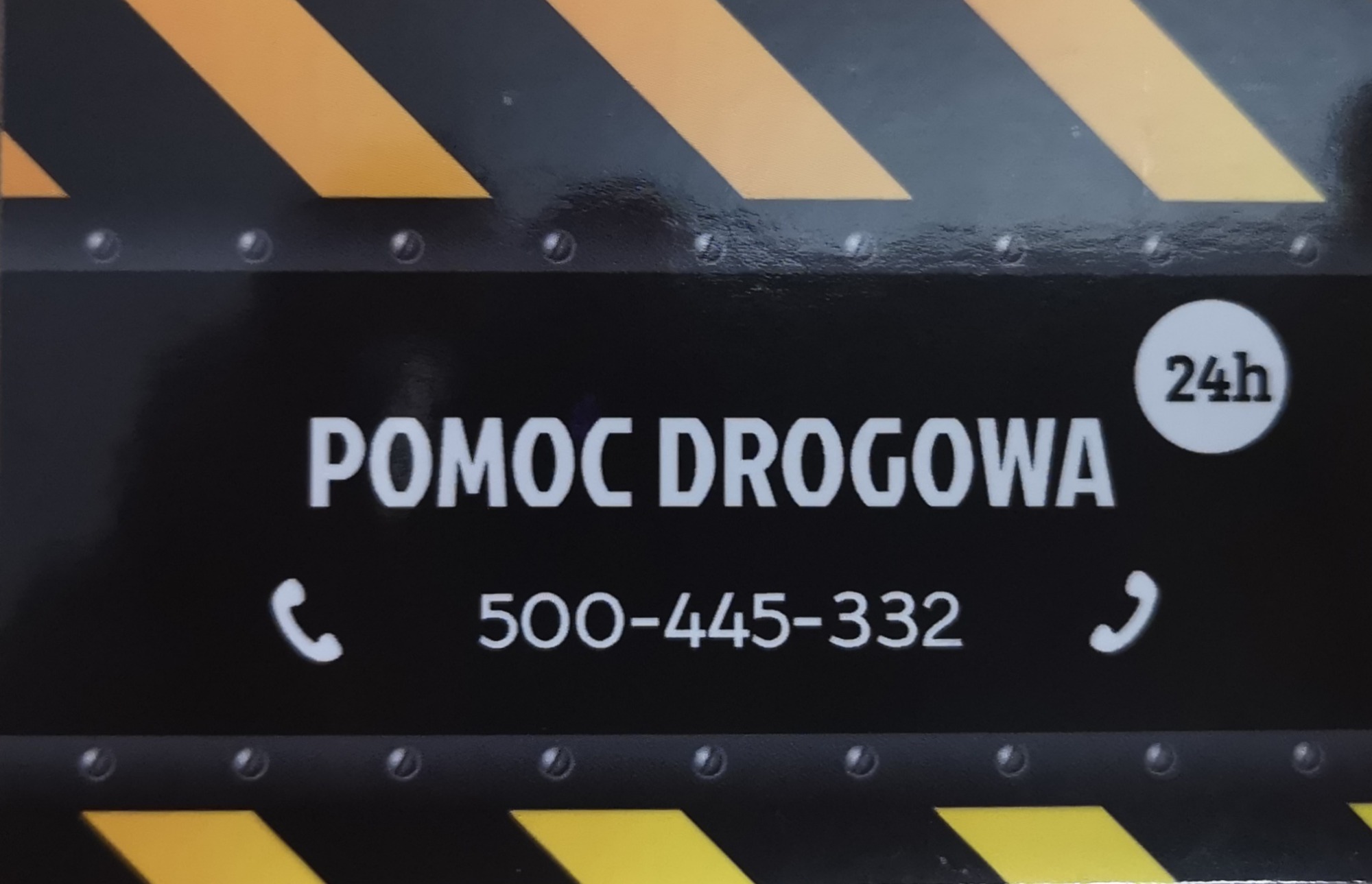 Czarno-żółta tabliczka z napisem 'POMOC DROGOWA' i numerem telefonu 500-445-332, informująca o dostępności 24h.