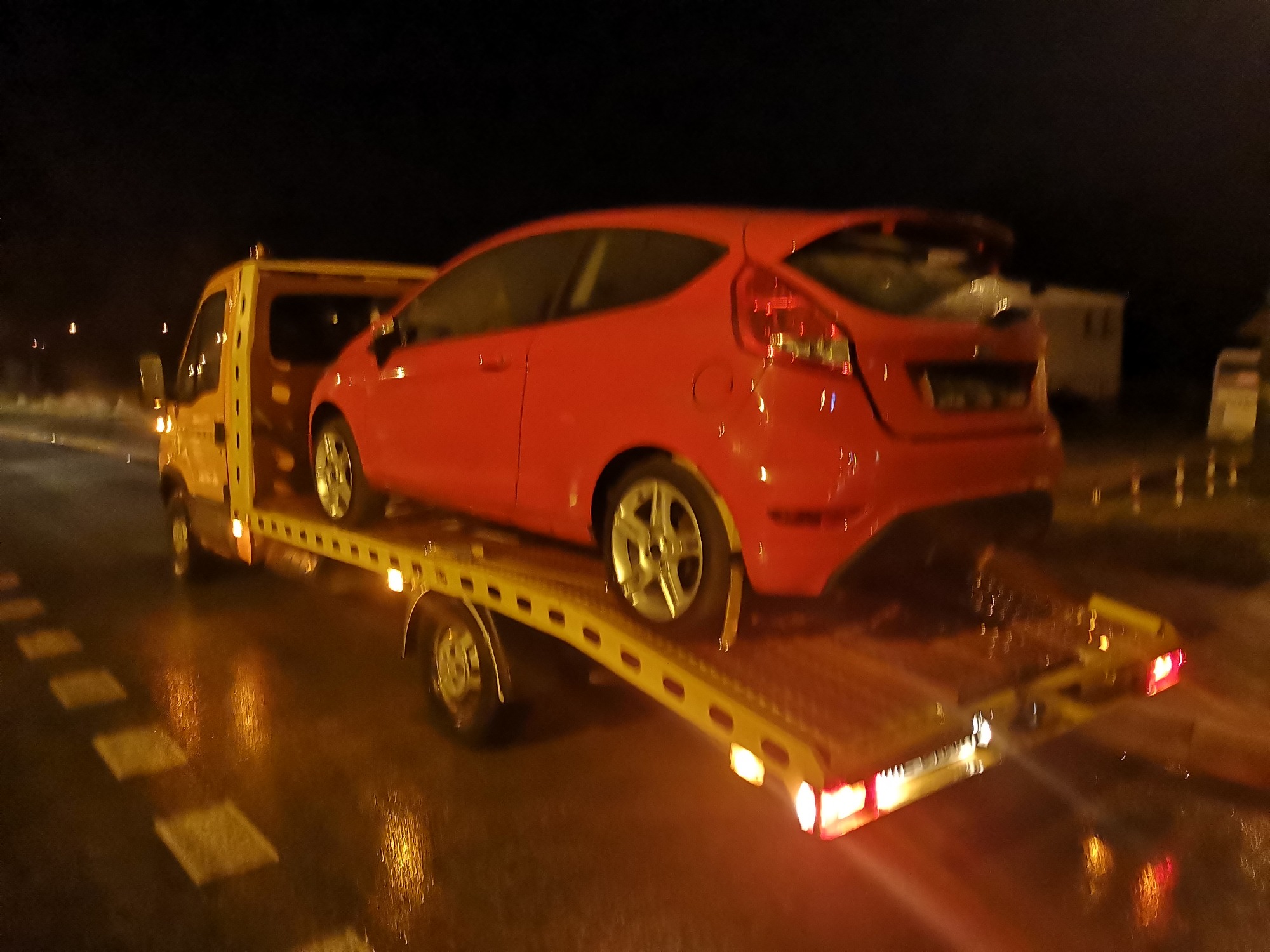Czerwony samochód osobowy Ford Fiesta na żółtej lawecie podczas nocnego transportu na mokrej jezdni.