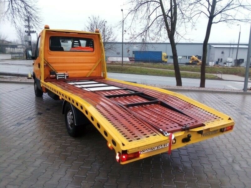 Żółta autolaweta z czerwoną platformą, widok z tyłu na mokrym bruku, zaparkowana na tle budynku przemysłowego i drzew bez liści.