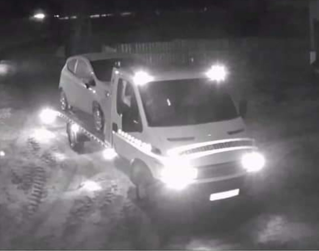 Nocne ujęcie lawety z załadowanym, jasnym samochodem osobowym na platformie, oświetlonej mocnymi reflektorami, widoczne ślady opon na podłożu.