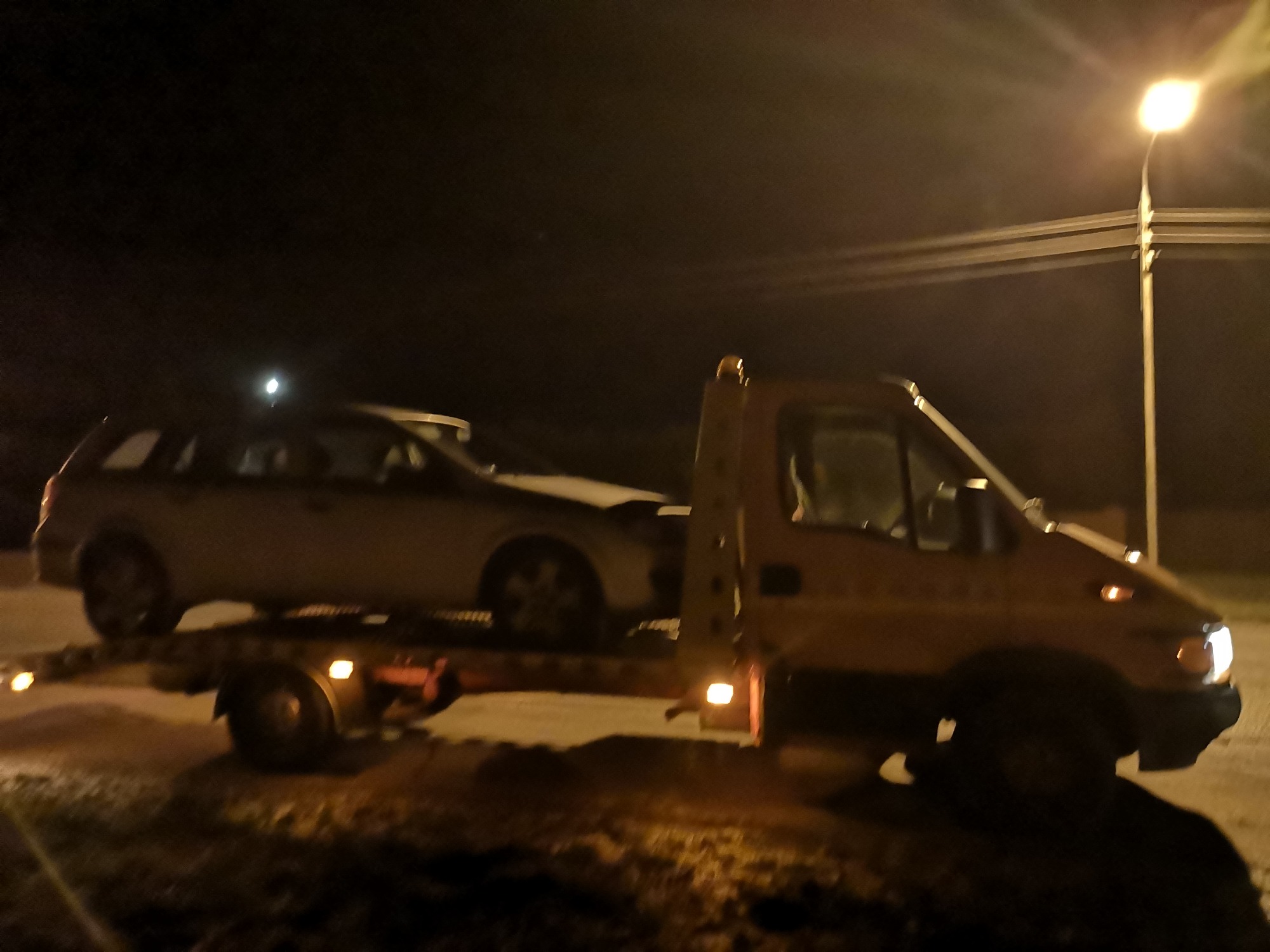 Nocny transport uszkodzonego kombi na platformie lawety, oświetlonej światłem latarni.