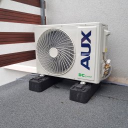 P.H.U. Eco-Constans - Biała jednostka zewnętrzna klimatyzacji AUX DC Inverter, zamontowana na dachu pokrytym szarą papą, na czarnych podkładkach antywibracyjnych, przy drewnianej balustradzie.