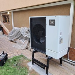 P.H.U. Eco-Constans - Biała pompa ciepła Mitsubishi Zubadan R32 na czarnym stelażu, zamontowana przy beżowej ścianie budynku z widocznym oknem i zielonym trawnikiem.