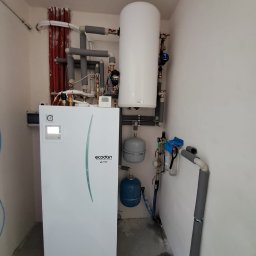 P.H.U. Eco-Constans - Biała pompa ciepła Ecodan Mitsubishi z zbiornikiem, rurami i osprzętem w niewielkim pomieszczeniu. Widoczne izolowane rury i elementy instalacji.