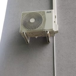 P.H.U. Eco-Constans - Klimatyzator AUX zamontowany na szarej, teksturowanej ścianie budynku z widoczną instalacją odprowadzającą. Liście drzew w górnym rogu kadru.