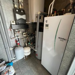 P.H.U. Eco-Constans - Kompletna instalacja pompy ciepła Ecodan Mitsubishi, z widocznymi rurami, zbiornikiem i modułem sterującym w kotłowni. Szare rury i biała obudowa pompy.