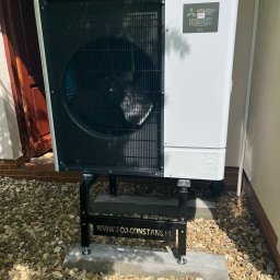 P.H.U. Eco-Constans - Biała pompa ciepła Mitsubishi Zubadan R32, zainstalowana na zewnątrz budynku na betonowej podstawie, otoczona kamieniami ozdobnymi i zielenią.