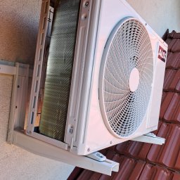 P.H.U. Eco-Constans - Biała jednostka zewnętrzna klimatyzatora AUX zamontowana na ścianie nad dachem z czerwonej dachówki, widoczne mocowania i fragment ściany z fakturą.