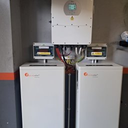 P.H.U. Eco-Constans - Dwa białe magazyny energii Felicitysolar z rozdzielnicami PV-DC i PV-AC oraz inwerter Deye na szarej ścianie z pomarańczowym pasem. Instalacja wewnątrz budynku.