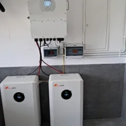 Biały inwerter Daye i dwa magazyny energii Felicity ESS zamontowane na ścianie w pomieszczeniu z szarą podłogą. Widoczne przewody i skrzynki elektryczne.