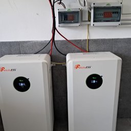 P.H.U. Eco-Constans - Dwa białe magazyny energii Felicity ESS z wyświetlaczami, zamontowane na tle szarej ściany z płytkami, z widocznymi przewodami i skrzynkami elektrycznymi powyżej.