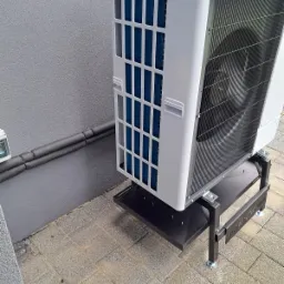 Zewnętrzna jednostka pompy ciepła na czarnym, metalowym stojaku, z widocznymi niebieskimi lamelami i szarą elewacją budynku w tle. Rury instalacyjne na ścianie.