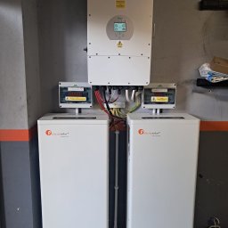 P.H.U. Eco-Constans - Dwa białe magazyny energii Felicitysolar z rozdzielnicami PV-AC/DC i falownikiem Deye na szarej, częściowo pomalowanej ścianie w garażu. Instalacja wewnątrz budynku.