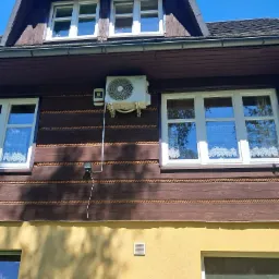 Biała klimatyzacja na drewnianej elewacji domu z brązowymi zdobieniami, okna z białymi ramami i firankami, błękitne niebo w tle.