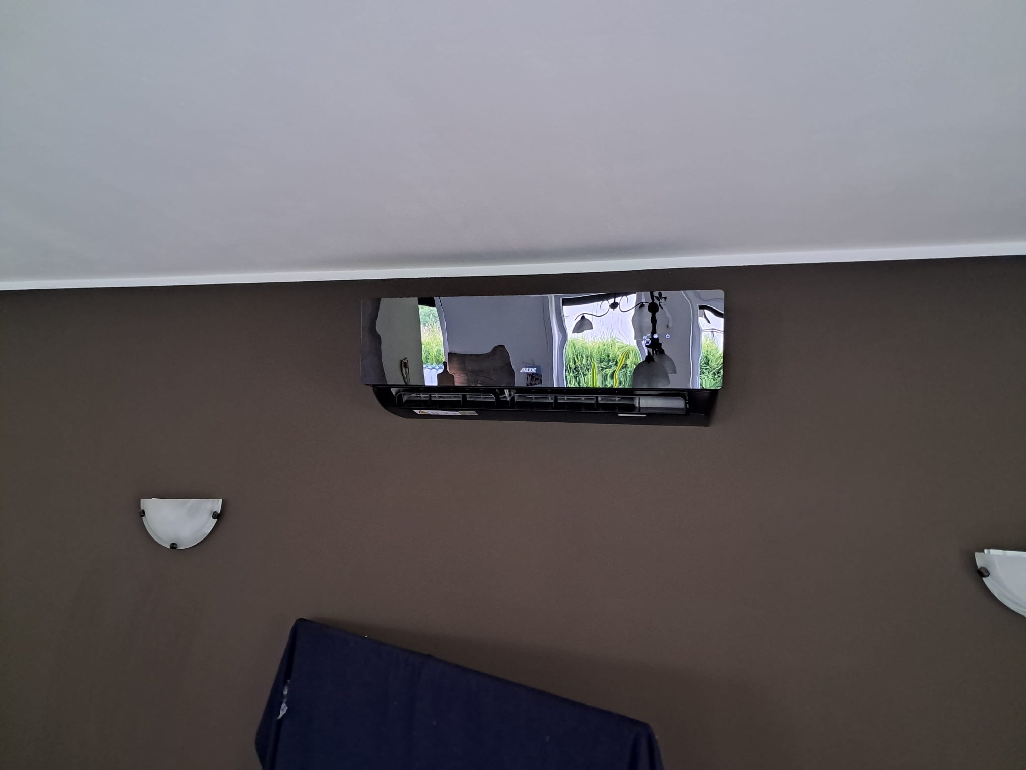 Czarny klimatyzator AUX zamontowany na brązowej ścianie w salonie. Widoczne lampy ścienne i odbicie wnętrza w obudowie klimatyzatora.