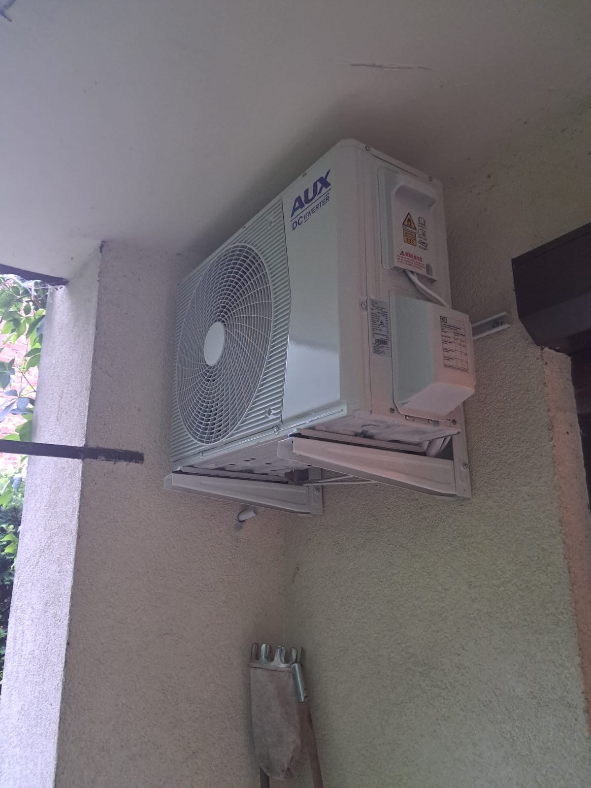 Biała jednostka zewnętrzna klimatyzacji AUX DC Inverter zamontowana na ścianie budynku o jasnej elewacji. Widoczne elementy montażowe i fragment ściany z fakturą.