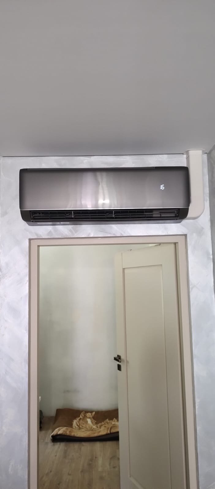 Czarny klimatyzator ścienny z wyświetlaczem temperatury, zamontowany nad drzwiami w pokoju z dekoracyjną ścianą. Widoczna część legowiska dla zwierząt w tle.