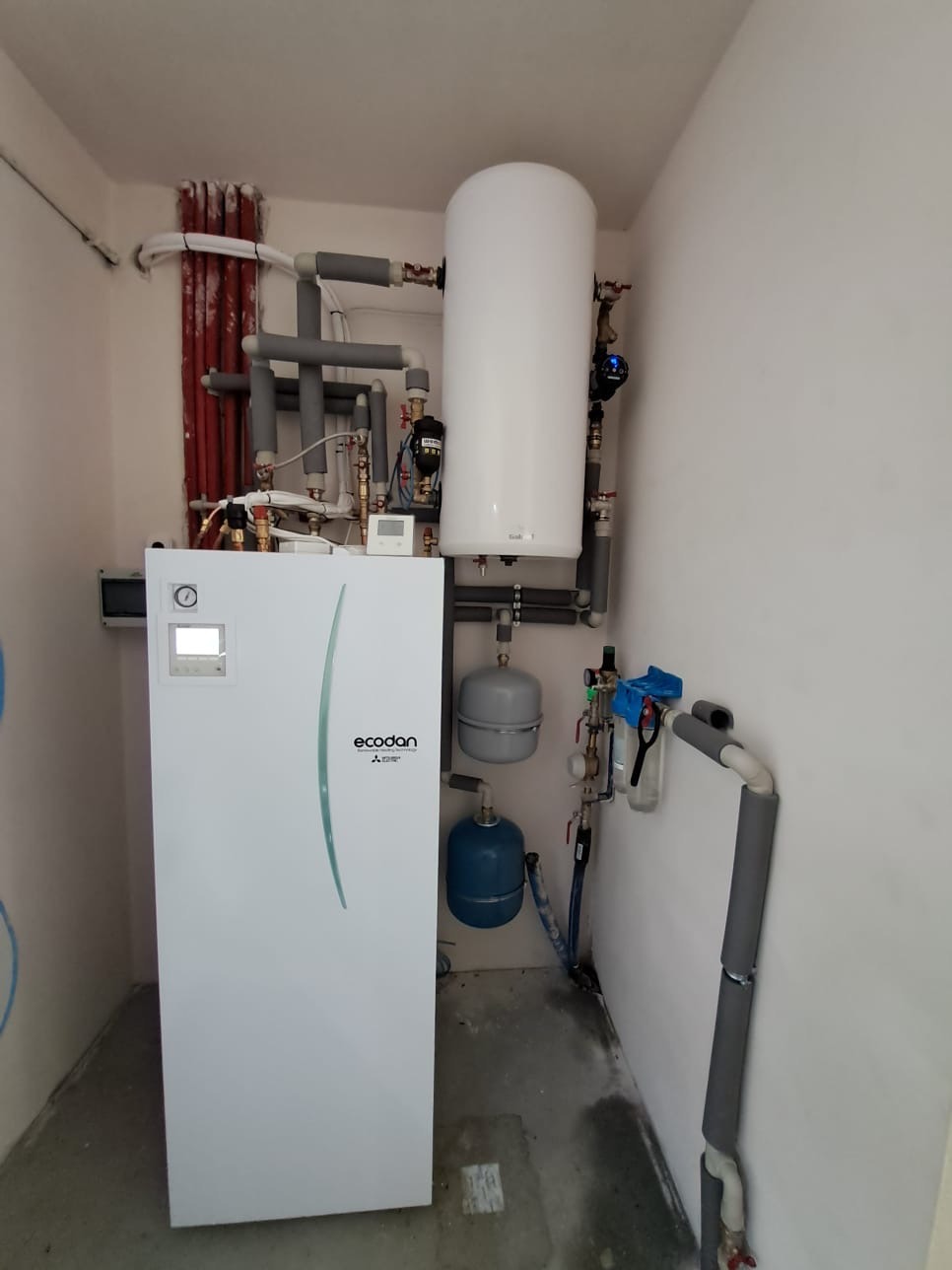 Biała pompa ciepła Ecodan Mitsubishi z zbiornikiem, rurami i osprzętem w niewielkim pomieszczeniu. Widoczne izolowane rury i elementy instalacji.