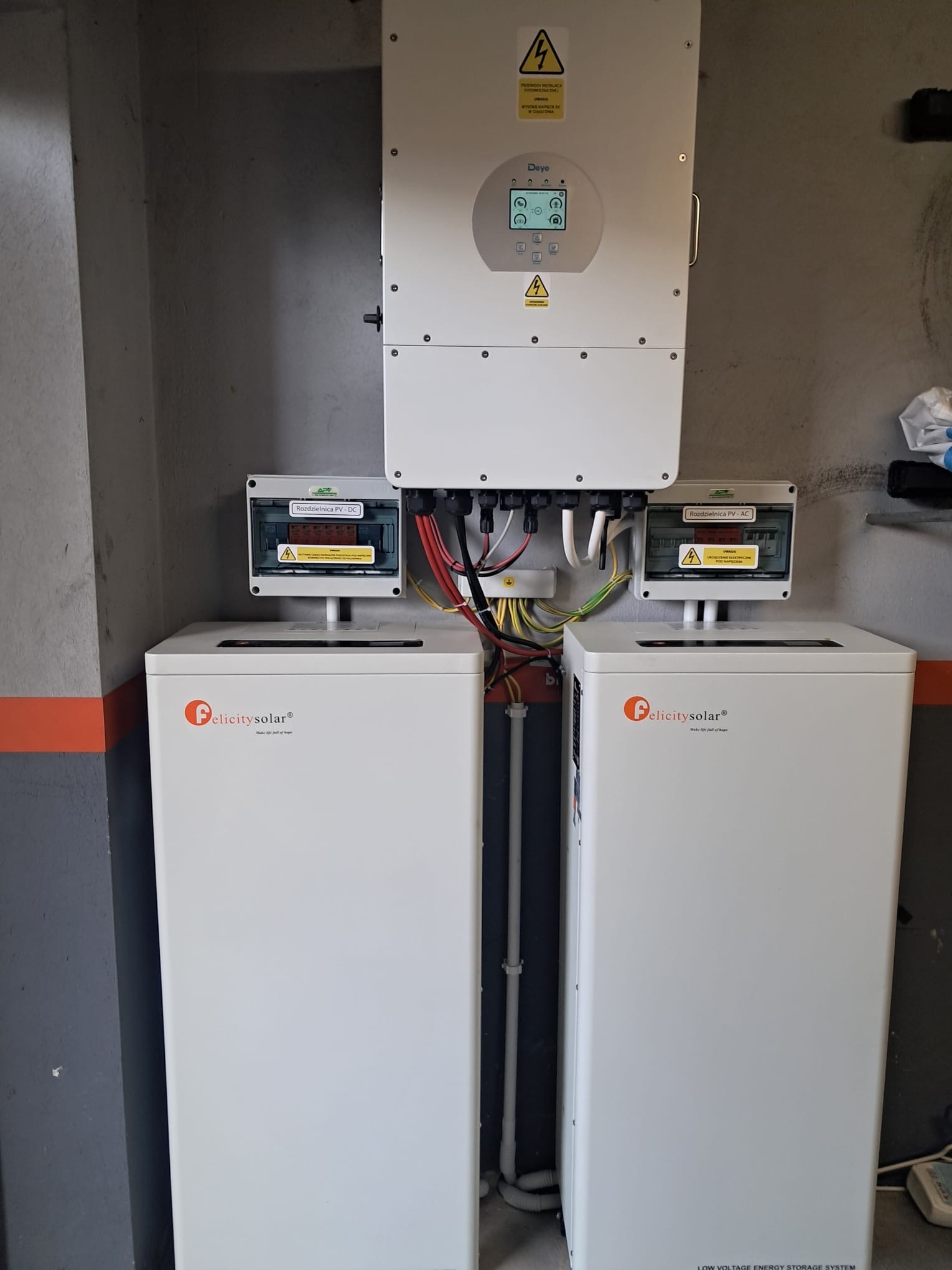 Dwa białe magazyny energii Felicitysolar z rozdzielnicami PV-DC i PV-AC oraz inwerter Deye na szarej ścianie z pomarańczowym pasem. Instalacja wewnątrz budynku.