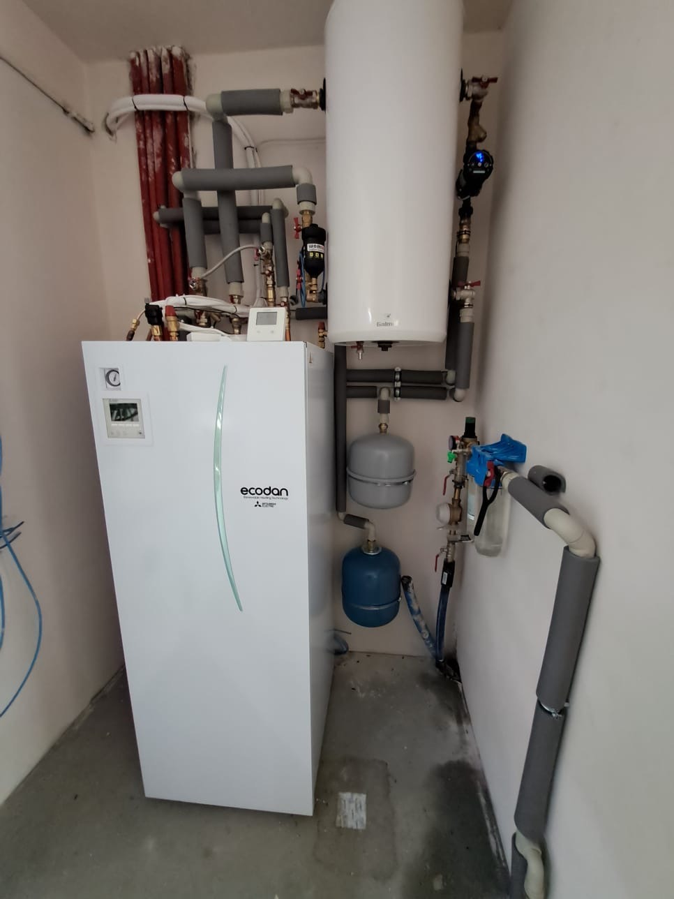 Biała pompa ciepła Ecodan Mitsubishi Electric z szarymi rurami w kotłowni. Widoczne zbiorniki wyrównawcze i osprzęt instalacyjny. Ujęcie z dołu, lekko z lewej.