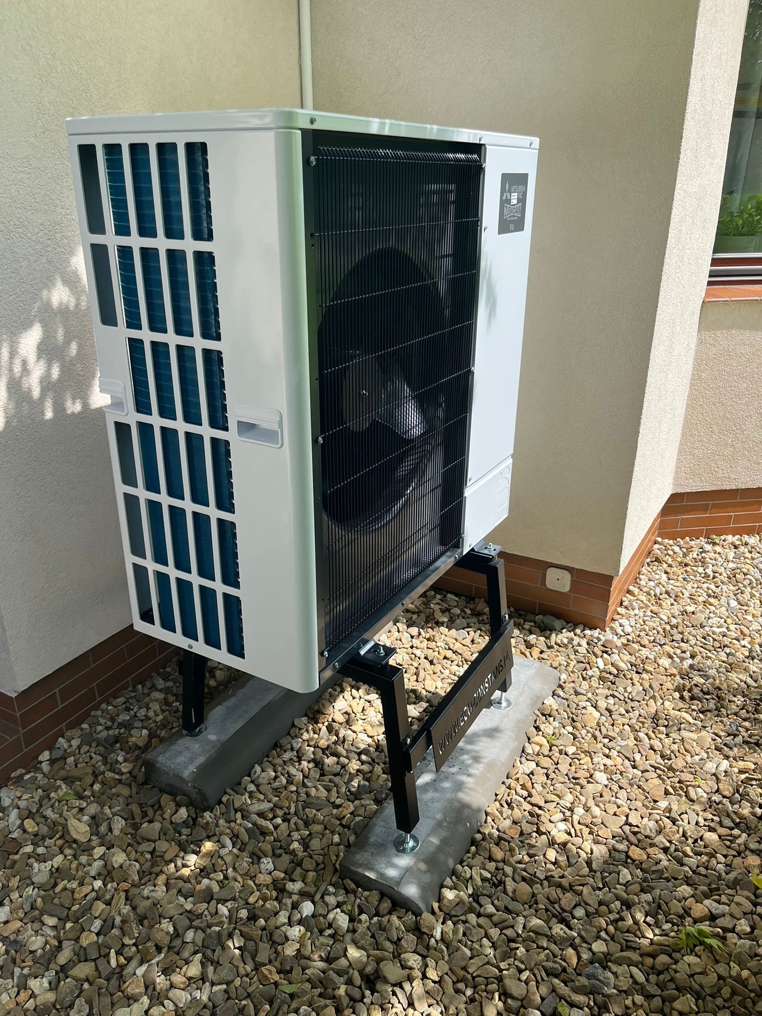 Biała pompa ciepła Mitsubishi Zubadan na czarnym stelażu, ustawiona na betonowych płytach na żwirowym podłożu przy ścianie budynku. Widoczne detale konstrukcyjne.