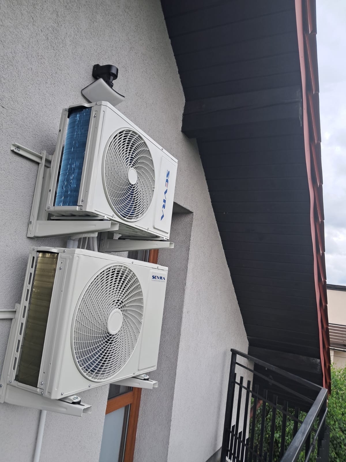 Dwa białe klimatyzatory marki Sevra zamontowane jeden nad drugim na szarej ścianie budynku z widocznym fragmentem dachu i balkonu.
