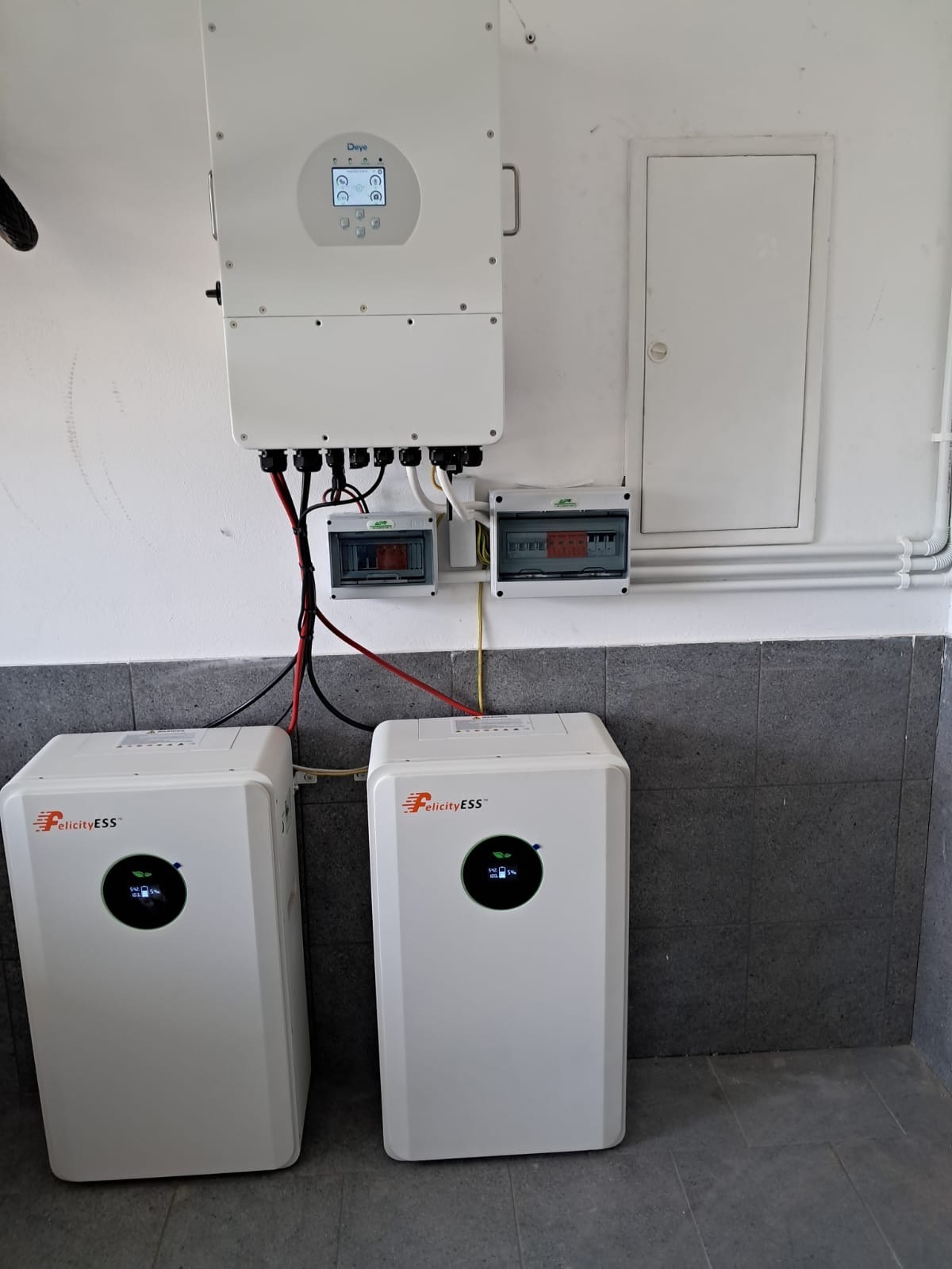 Biały inwerter Daye i dwa magazyny energii Felicity ESS zamontowane na ścianie w pomieszczeniu z szarą podłogą. Widoczne przewody i skrzynki elektryczne.