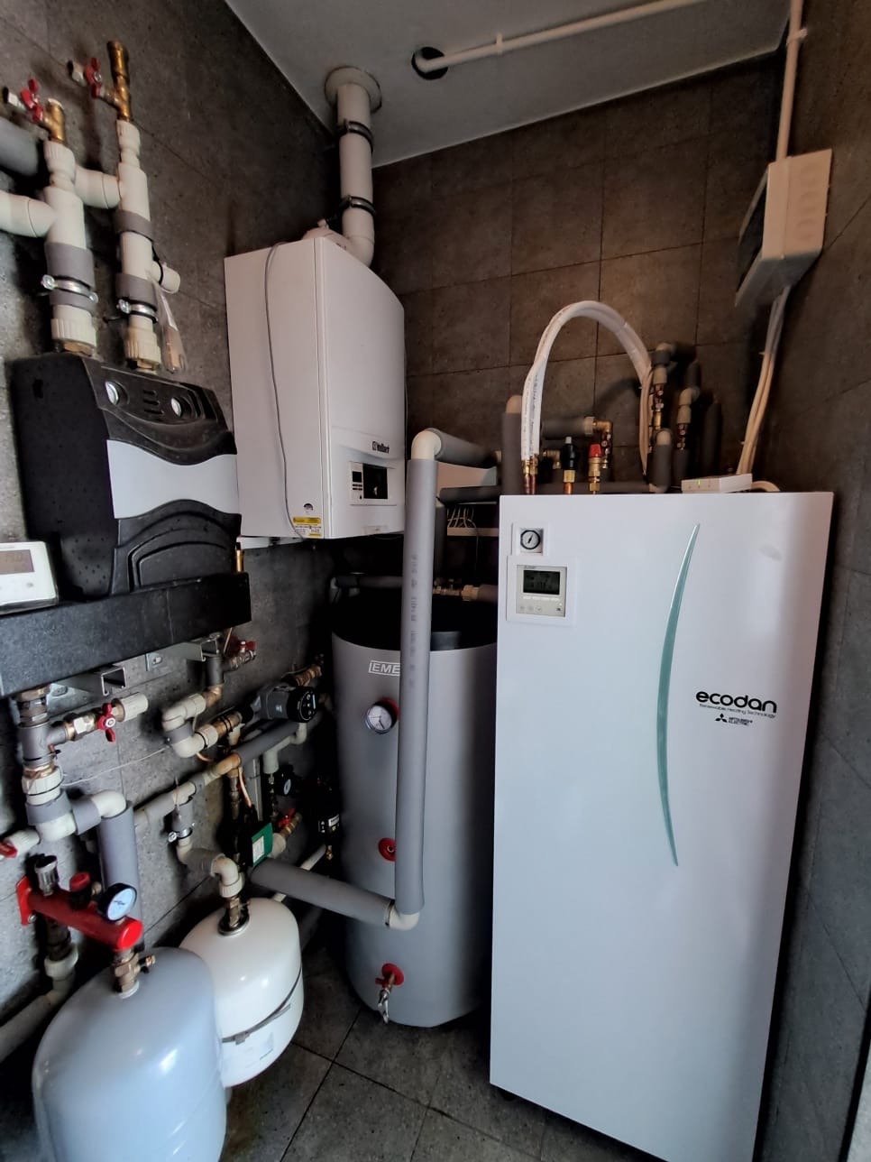 Kompletna instalacja pompy ciepła Ecodan Mitsubishi z zasobnikiem i kotłem Vaillant w kotłowni z szarymi płytkami. Widoczne rury i zawory.