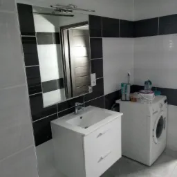 Wykończona łazienka z białą szafką pod umywalkę, lustrem z czarno-białymi płytkami i pralką, z widocznymi akcesoriami kosmetycznymi na pralce.