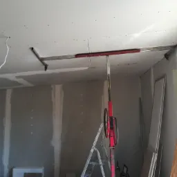 Montaż płyty gipsowo-kartonowej na suficie za pomocą podnośnika, widoczne profile i wstępnie zamocowane płyty, ściany w stanie surowym.