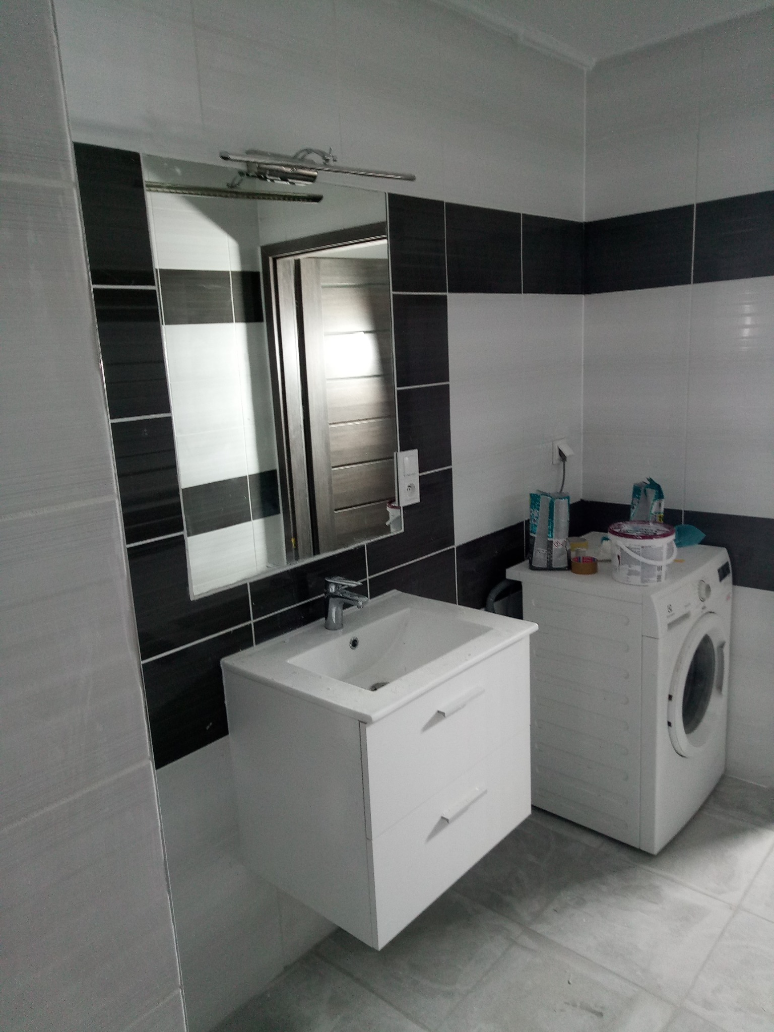 Wykończona łazienka z białą szafką pod umywalkę, lustrem z czarno-białymi płytkami i pralką, z widocznymi akcesoriami kosmetycznymi na pralce.
